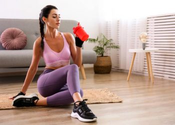 Stay at home training concept. Young attractive woman of athletic build drinking post workout protein shake. Beautiful female in feminine sportswear holding red shaker. Close up, copy space background